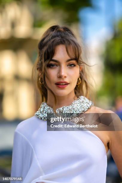 Mara Lafontan wears gold earrings, a rhinestones large necklace, a white asymmetric shoulder / long train sleeves top, outside Stéphane Rolland,...