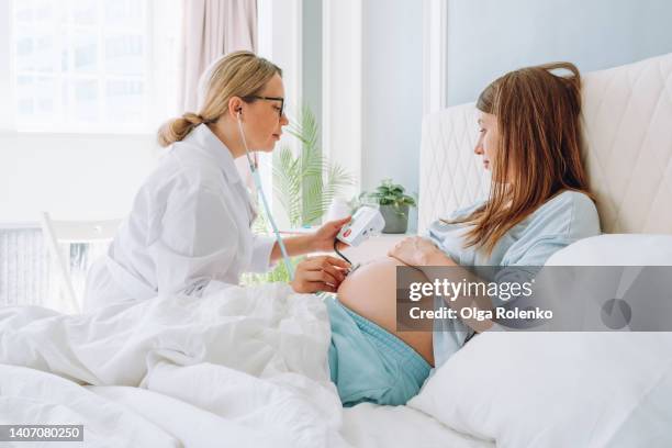 female doctor in glasses listening to belly of pregnant woman lying on the couch, using stethoscope - herzton abhören stock-fotos und bilder