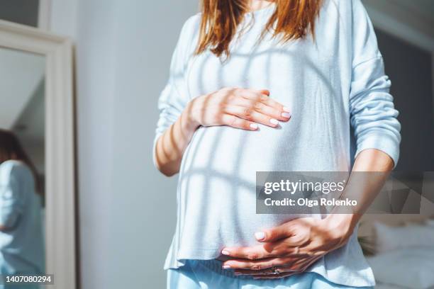 close up cropped pregnant woman holding her abdomen with love, care of unborn baby and standing near mirror - holprig stock-fotos und bilder