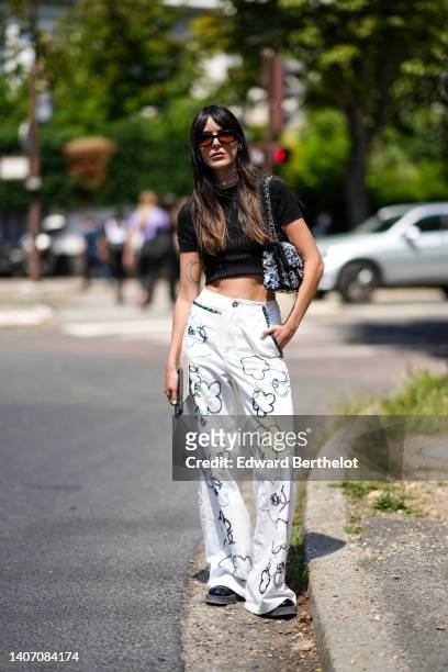 Leia Sfez wears black sunglasses, silver earrings, a silver and diamonds flower pattern necklace, a black short sleeves / cropped t-shirt from...