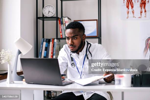 serious african-american man doctor is wearing a lab coat and a stethoscope using laptop in doctor's office. - computer lab stock-fotos und bilder