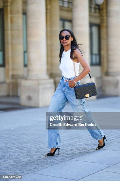 Emilie Joseph wears black sunglasses, a white halter-neck tank-top, a black shiny grained leather Boy shoulder bag from Chanel, blue denim cut-out...