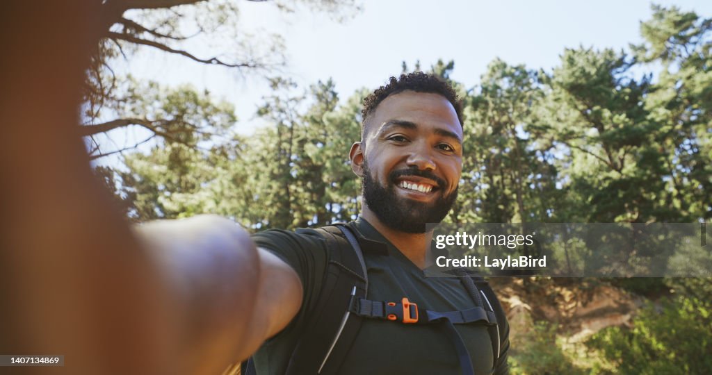 Active fit african american male taking a selfie photo and smiling while out on a hike in a natural green forest in the morning on a summer day. Healthy hiker enjoying the beauty of outdoor nature