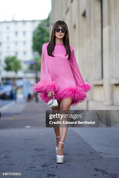 Guest wears black sunglasses, a neon pink oversized long sleeves / short dress with feathers borders, a white nailed / studded handbag from...