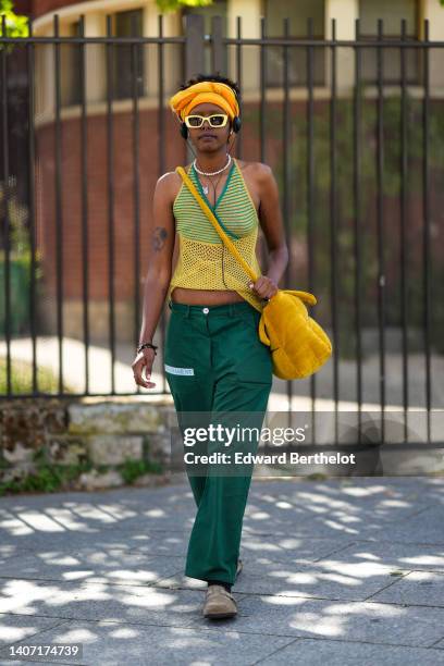 Guest wears a yellow scarf as a headband, yellow sunglasses, black headphones, a silver piercing, a white pearls necklace, a yellow and green striped...