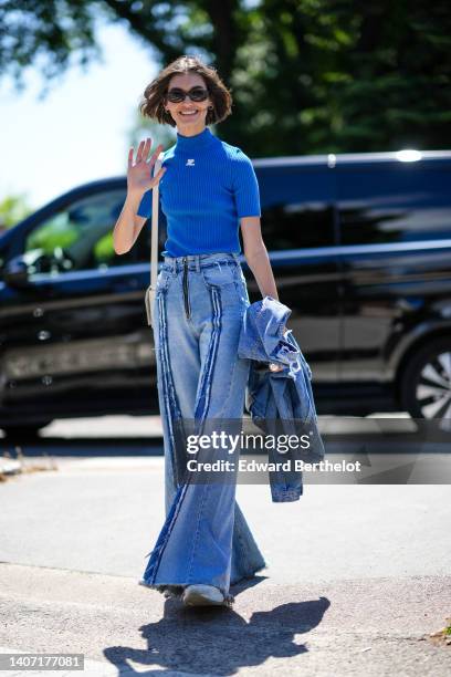 Model wears black sunglasses, gold earrings, a blue turtleneck / short sleeves with embroidered white logo t-shirt from Courreges, a white leather...