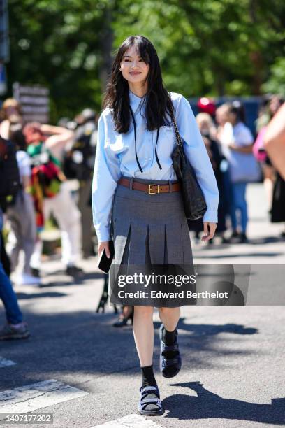 Model wears a baby blue shirt, a brown shiny leather belt, a black shiny leather Chanel 22 handbag from Chanel, a dark gray pleated / accordion knees...