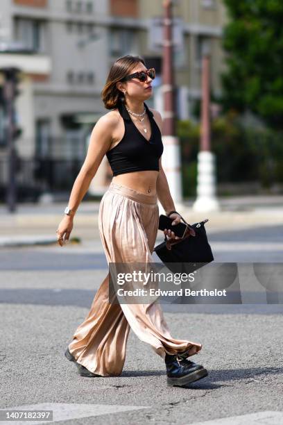 Guest wears sunglasses, gold earrings, a white pearls necklace, gold long pendant necklace, a black ribbed V-neck cropped top, beige silk flowing...