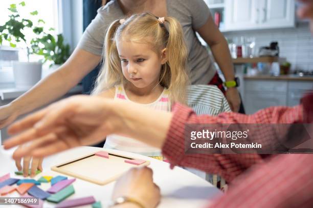 little serious 5-years old blond girl playing wooden puzzle with her mother and grandmother at dinner table at home. - 50 54 years stockfoto's en -beelden