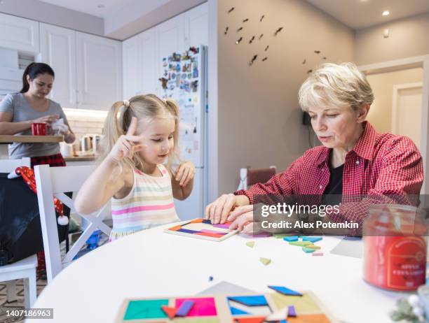 la bambina concentrata gioca con il puzzle tangram a tavola in cucina con sua nonna. - tangram foto e immagini stock