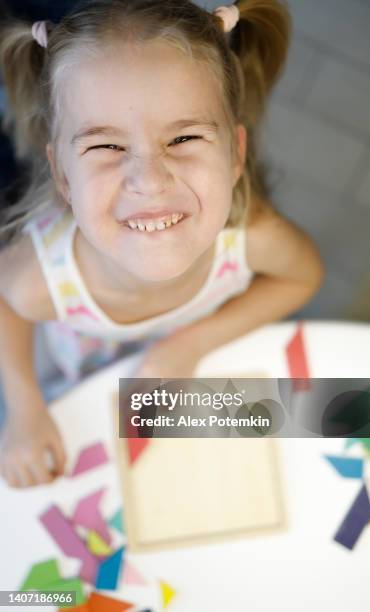 ritratto di bambina bionda sorridente che gioca con il puzzle tangram a casa. direttamente sopra la vista con messa a fuoco selettiva sul suo viso e sfondo sfocato. - tangram foto e immagini stock