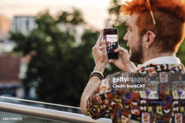 rear view on a young alternative-looking gay man using smart phone and photographing sunset - cat hipster no stock pictures, royalty-free photos & images