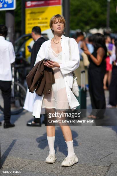 Model wears gold earrings, a gold chain necklace, a white lace V-neck transparent short dress, a white oversized shirt, a brown jacket, a white...
