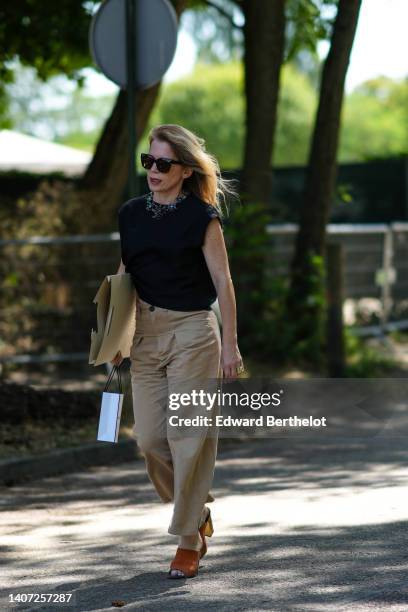 Guest wears black sunglasses, a silver large necklace, a black short sleeves t-shirt, beige large pants, camel shiny leather open toe-cap / block...