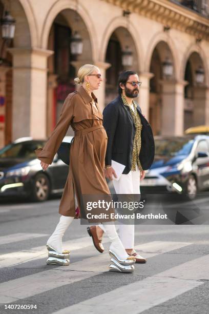 Guest wears cat eyes sunglasses, gold large earrings from Schiaparelli, a brown belted long trench coat, white leather open toe-cap / oversized...