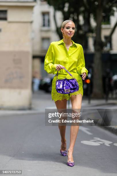 Guest wears gold earrings, a yellow silk shirt, matching yellow lace pattern shorts, a purple shiny sequined handbag from Fendi, a gold large pendant...