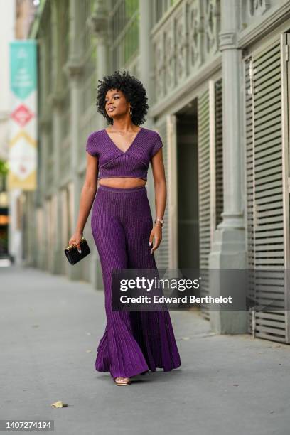 Didi Stone wears gold earrings, a gold and diamonds necklace from Messika, a purple ribbed short sleeves / V-neck cropped top, matching purple and...