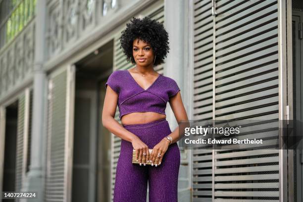Didi Stone wears gold earrings, a gold and diamonds necklace from Messika, a purple ribbed short sleeves / V-neck cropped top, matching purple and...