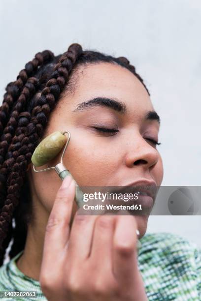 young woman with eyes closed using jade roller against white background - 玉石滾輪 個照片及圖片檔