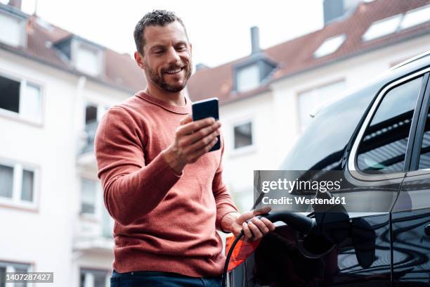 smiling man using smart phone charging electric car outside building - using phone in car stock pictures, royalty-free photos & images