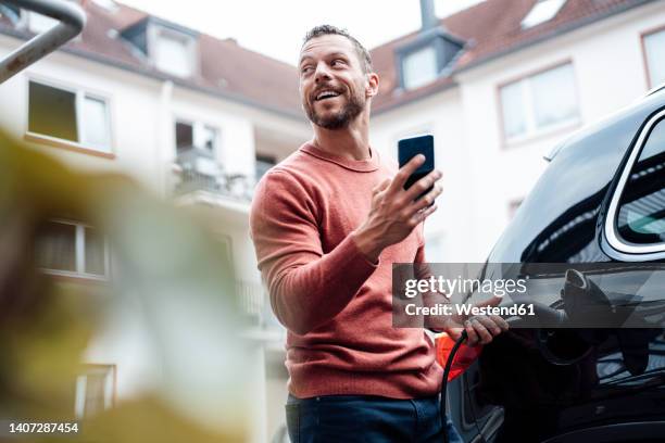 happy man with smart phone charging electric car at station - guy in car stock-fotos und bilder