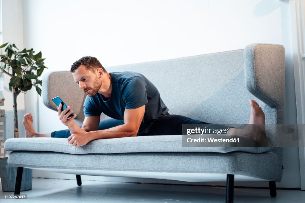 Man using smart phone sitting with legs apart on sofa at home