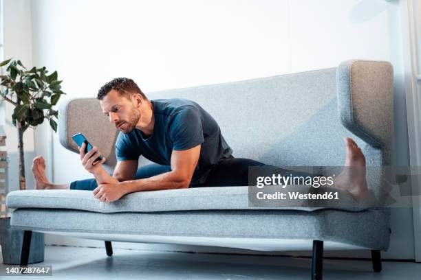 man using smart phone sitting with legs apart on sofa at home - legs spread - fotografias e filmes do acervo
