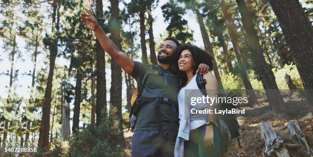 happy couple hiking a trail and enjoying the view in a forest. cheerful adventurous man and woman pointing at scenery while taking a break from exploring their surroundings during a trek in nature - young couple hiking stock pictures, royalty-free photos & images