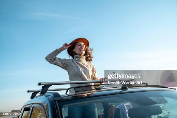 cheerful young woman wearing hat enjoying through sun roof of car - car sunroof stockfoto's en -beelden