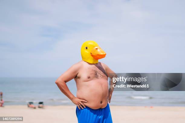 shirtless obese man with hand on hips wearing duck mask standing at beach - fat guy on beach photos et images de collection