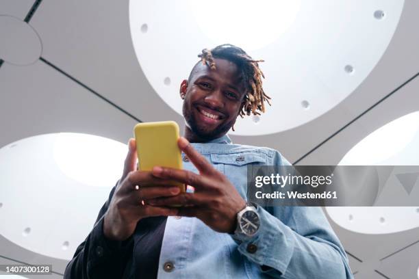 smiling man holding smart phone standing under illuminated ceiling - low angle view man stock pictures, royalty-free photos & images