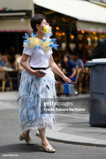 Guest wears diamond earrings, a yellow with embroidered white pattern face mask, a white t-shirt with embroidered blue and yellow ruffled flower...