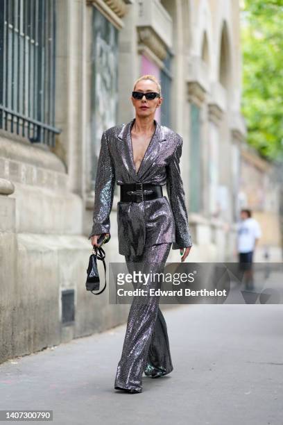 Guest wears black sunglasses, a silver sequined oversized blazer jacket, silver sequined flared pants, a black shiny leather large belt, black shiny...