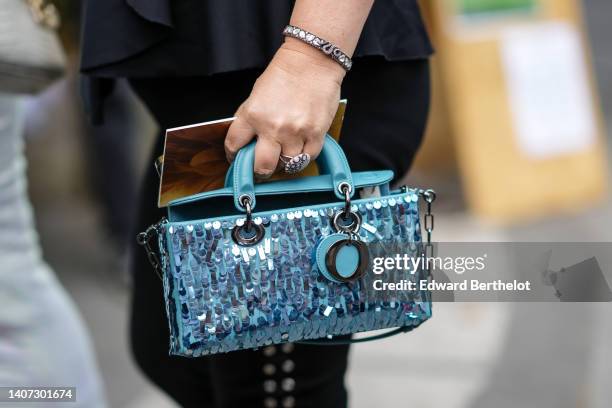 Guest wears a black ruffled V-neck / sleeveless t-shirt, silver bracelets, rings, a blue shiny leather with embroidered sequined handbag from Dior,...