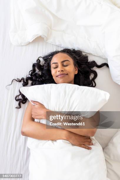 happy woman with eyes closed embracing cushion on bed in bedroom - man and woman cuddling in bed stock-fotos und bilder