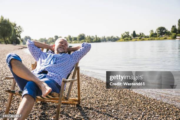 smiling man with hands behind head resting on deck chair at riverbank - beach deck chairs stock pictures, royalty-free photos & images