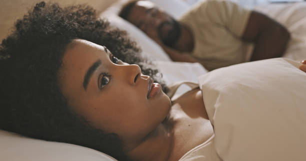 worried black woman laying in bed with insomnia looking anxious and concerned, having infidelity and relationship issues. man sleeping while his wife lays awake at night feeling depressed or troubled - black couple sleeping in bed stockfoto's en -beelden