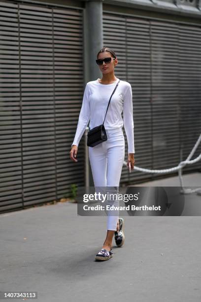 Guest wears black sunglasses, a white long sleeves t-shirt, a black shiny leather crocodile print pattern crossbody bag, white denim skinny pants,...