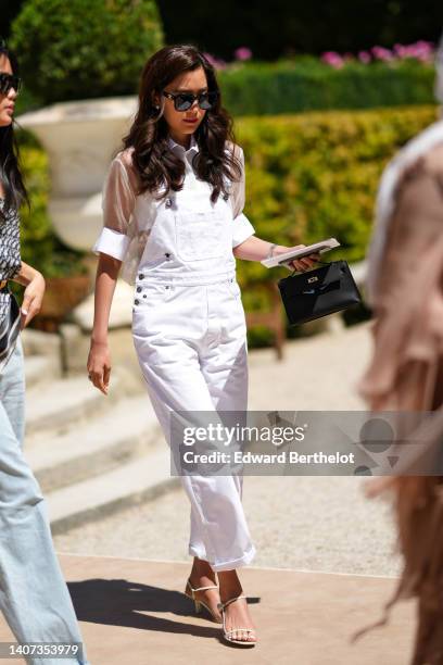 Guest wears black sunglasses from Celine, silver earrings, a white transparent short sleeves t-shirt, a white denim pair of overalls, a black shiny...