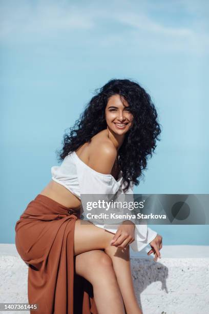 young beautiful mixed race woman with long curly black hair stands on the beach against the sky in a brown skirt and white blouse breathes the sea breeze - beautiful armenian women fotografías e imágenes de stock