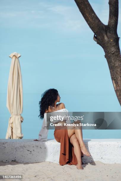 young beautiful mixed race woman with long curly black hair sits on a sandy beach in a brown skirt and white blouse, looks at the camera and smiles - beautiful armenian women fotografías e imágenes de stock