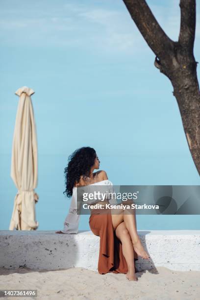 young beautiful mixed race woman with long curly black hair sits on a sandy beach in a brown skirt and white blouse, looks at the camera and smiles - beautiful armenian women stock pictures, royalty-free photos & images