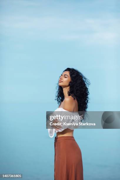 young beautiful mixed race woman with long curly black hair stands on the beach against the sky in a brown skirt and white blouse breathes the sea breeze - beautiful armenian women fotografías e imágenes de stock