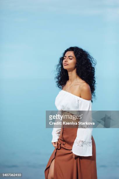 young beautiful mixed race woman with long curly black hair stands on the beach against the sky in a brown skirt and white blouse breathes the sea breeze - beautiful armenian women fotografías e imágenes de stock