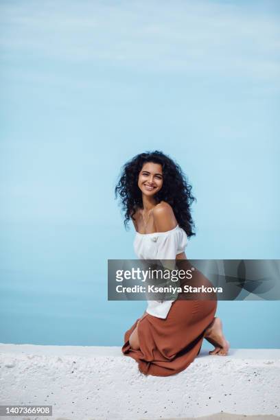 young beautiful mixed race woman with long curly black hair sits on a white pier in a brown skirt and white blouse, looks at the camera and smiles - beautiful armenian women fotografías e imágenes de stock