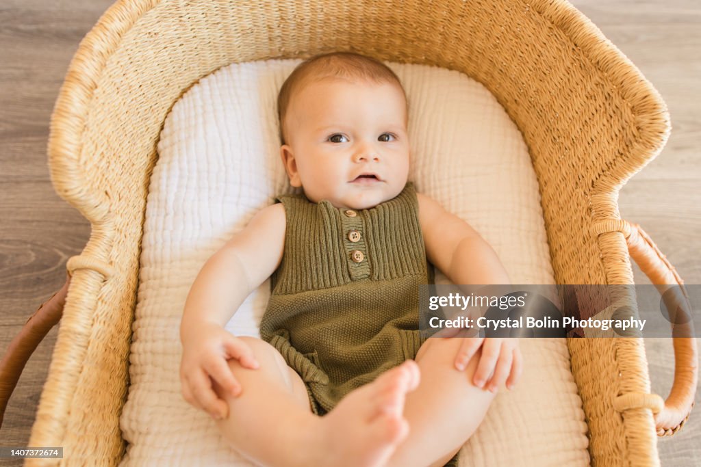 A 27-Week-Old Baby Boy Looking at the Camera With His Feet Up in the Air, Wearing an Olive Green Knit Romper While Laying in a Cozy Cotton Blanket in a Seagrass Moses Basket in a Neutral Modern Home
