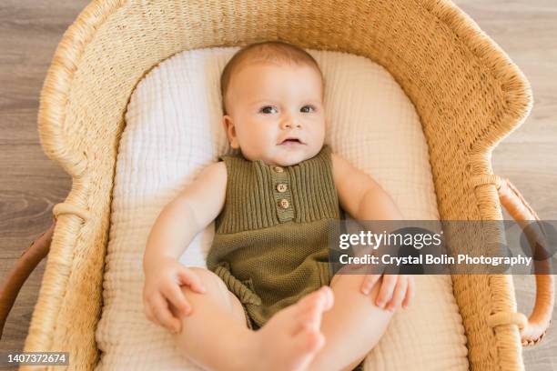 a 27-week-old baby boy looking at the camera with his feet up in the air, wearing an olive green knit romper while laying in a cozy cotton blanket in a seagrass moses basket in a neutral modern home - baby clothes stock pictures, royalty-free photos & images