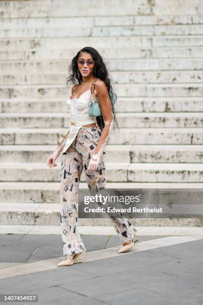 Winnie Harlow wears black sunglasses, gold and diamonds pendant earrings, a white latte shoulder-off / asymmetric corset top, a blue green matte...