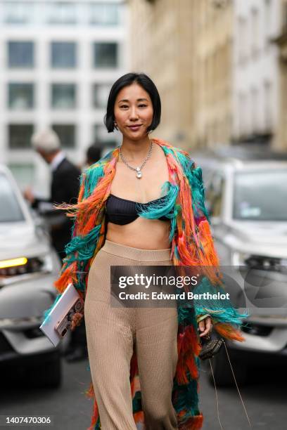 Guest wears earrings, a silver large chain pendant necklace, a black bra underwear, high waist beige ribbed flared pants, a green and orange print...