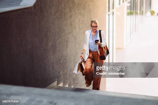 donna felice che tiene borse della spesa e usa il telefono cellulare all'aperto - vestito marrone foto e immagini stock
